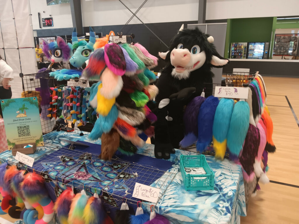 a photo of a cow mascot standing at a booth full of various tails, masks, paws, and other costume accessories in an auditorium. This photo is from Meadowlark Comic Con 2026 in Medford, OR